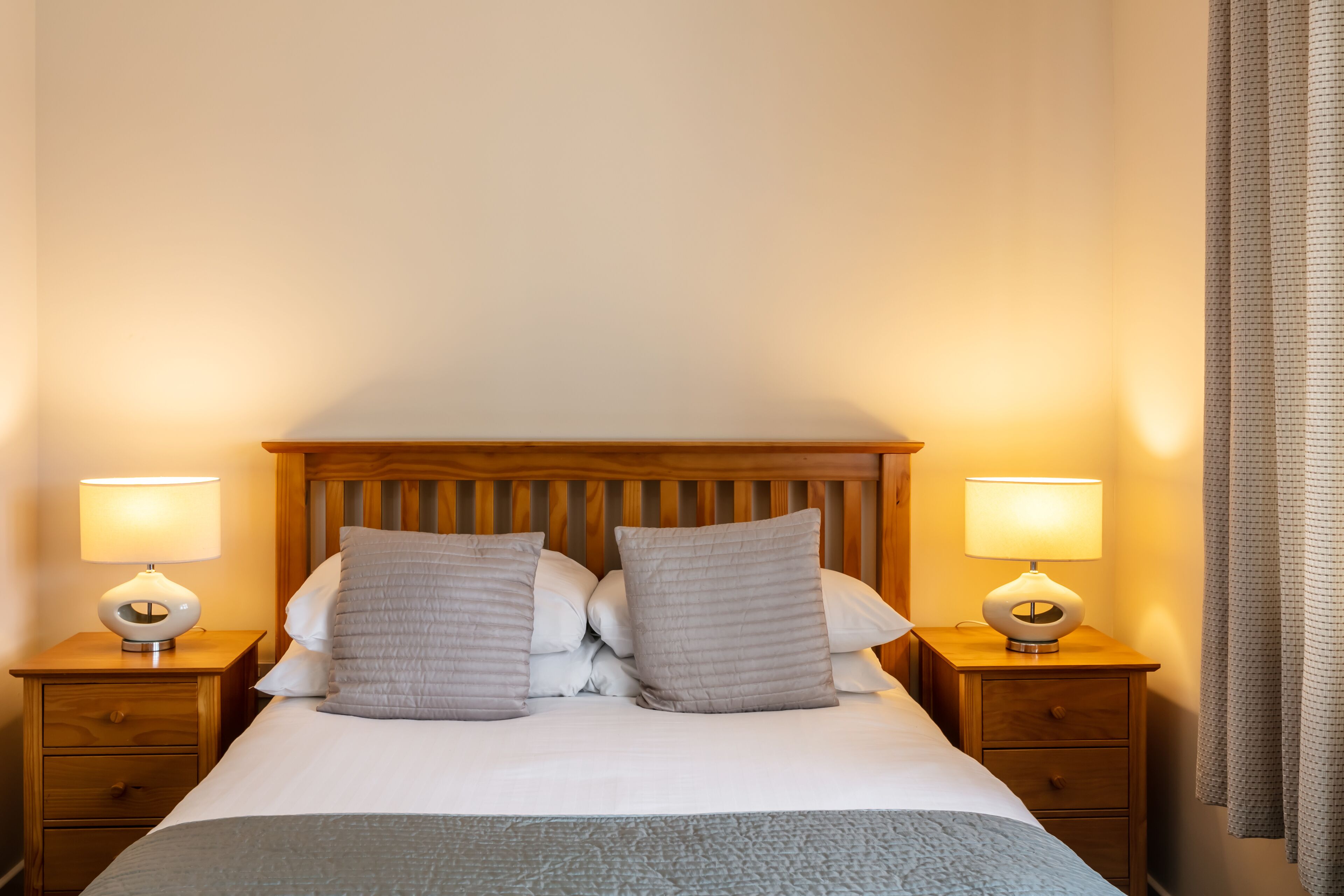 Een netjes opgemaakt bed met een houten hoofdbord, twee nachtkastjes met lampen en een grijze plaids.