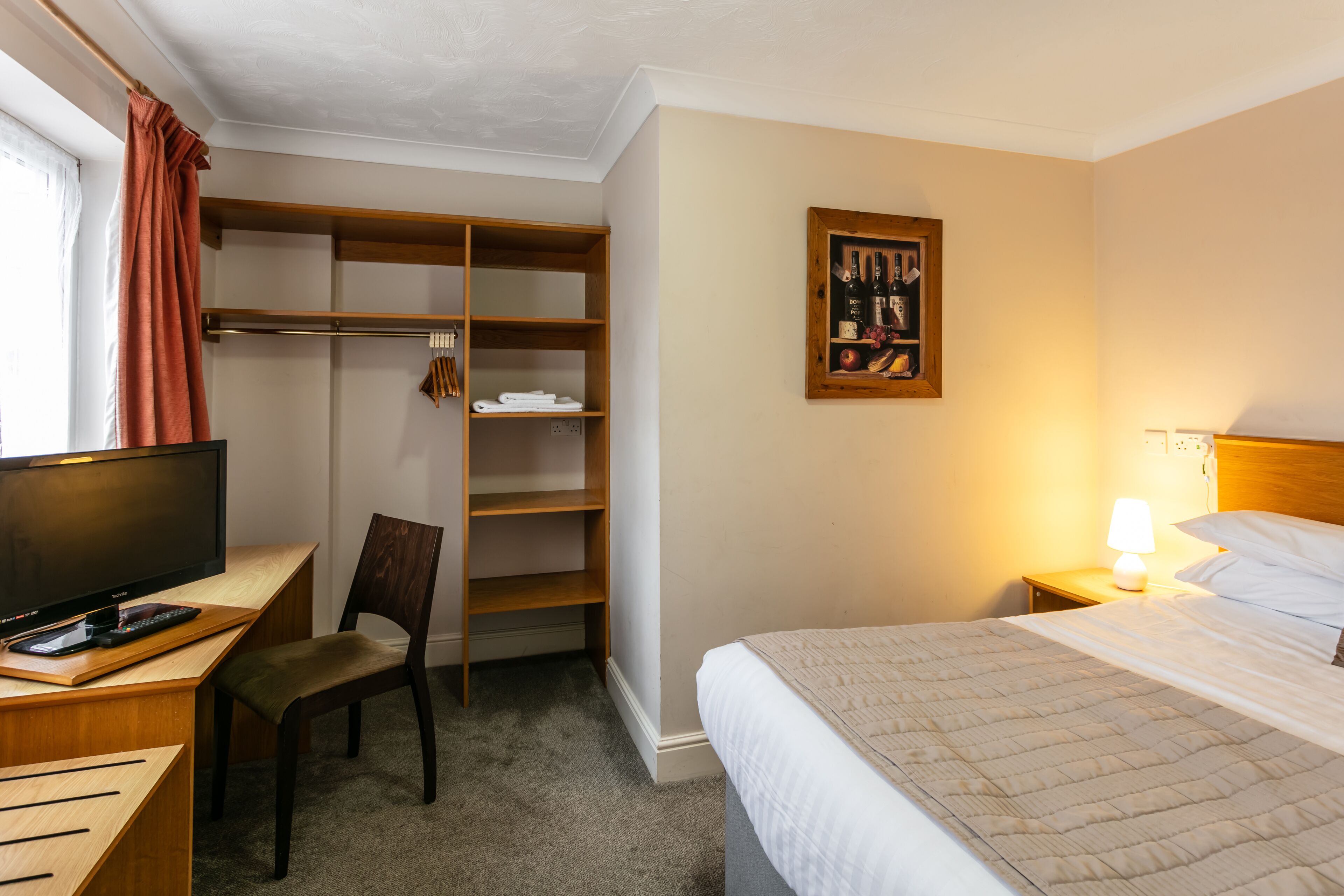 A hotel room with a bed, a desk with a computer, a chair, a wardrobe, and a wall-mounted painting.