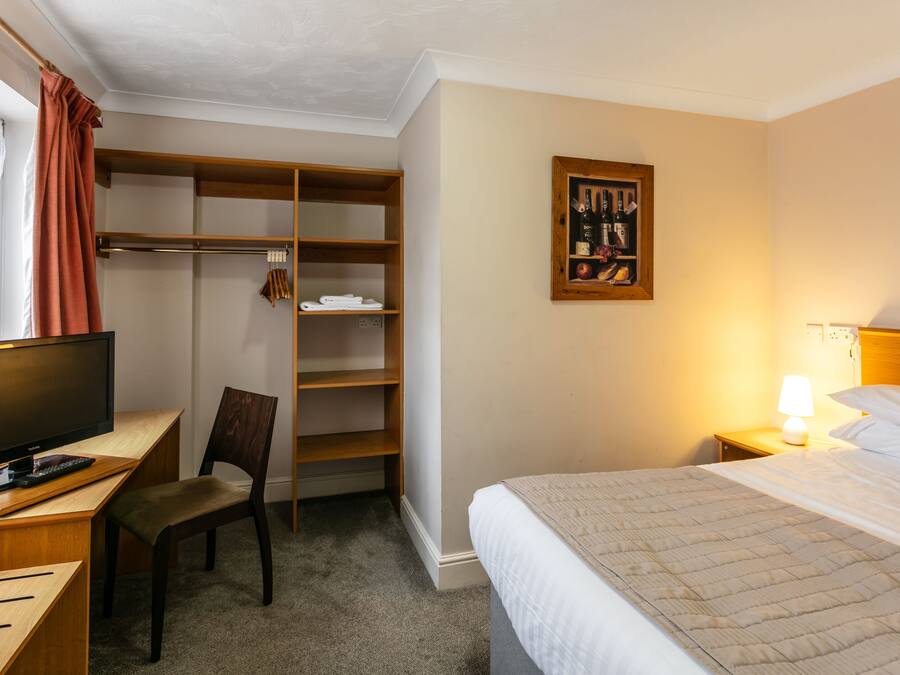 A hotel room with a bed, a desk with a computer, a chair, a wardrobe, and a wall-mounted painting.