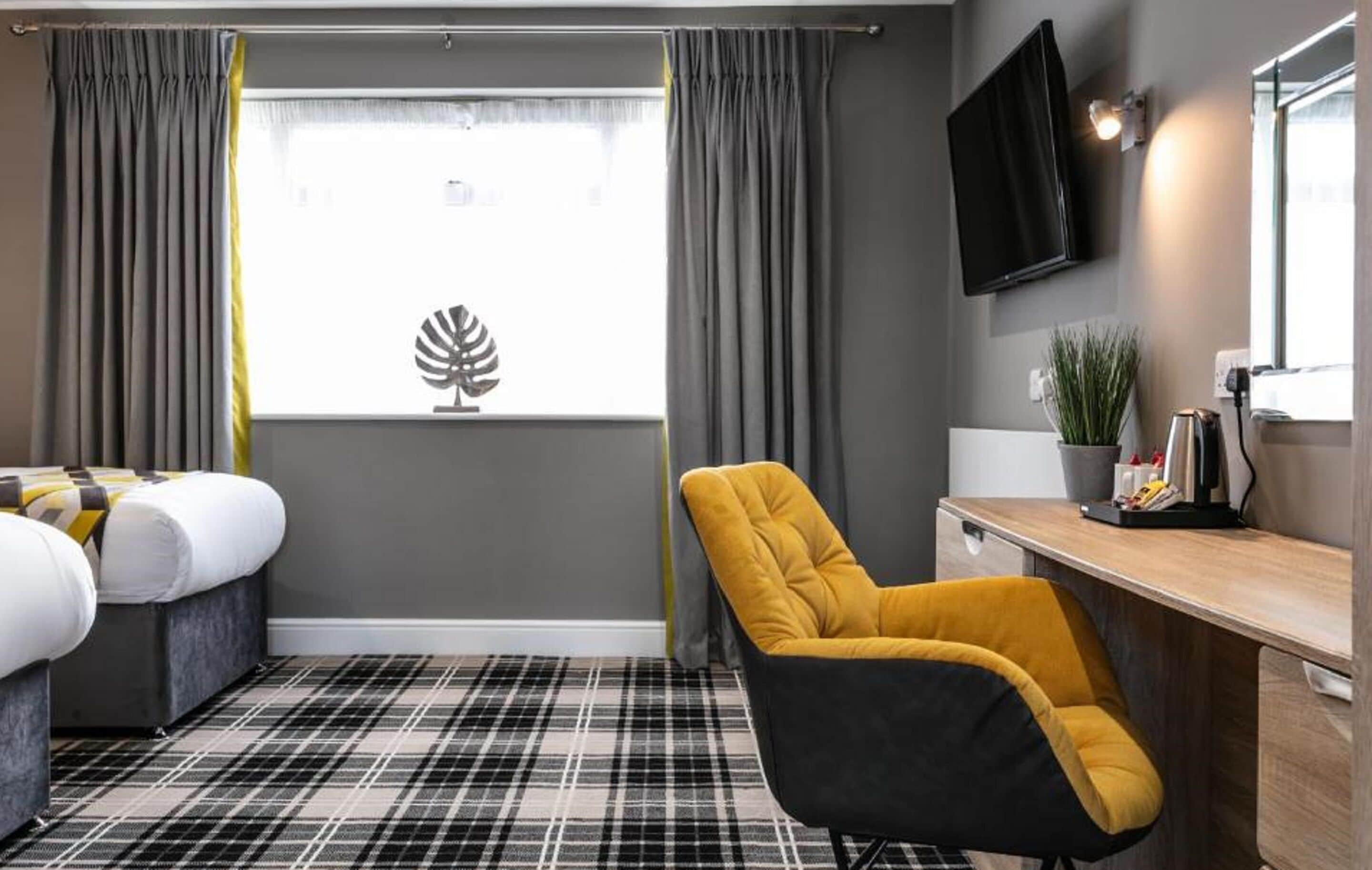 Une chambre d’hôtel avec un lit, un fauteuil jaune, un bureau avec une plante et un téléviseur à écran plat fixé au mur.