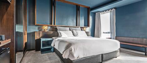 Una habitación de hotel moderna con una cama grande, una mesita de noche, un banco empotrado y vistas de la ciudad a través de la ventana.