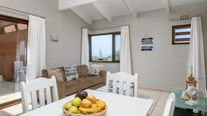 A dining area with a table set for two, a wicker fruit basket, a brown sofa with a cushion, and a window with a view of the sea. - San Lameer Villa  Rentals 2507 (Southbroom)