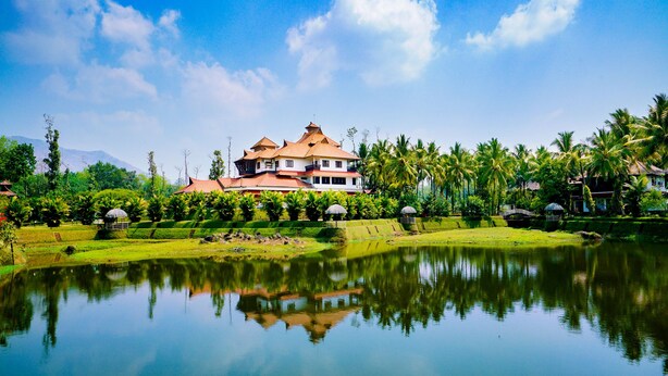 A large house with a red roof, surrounded by a pond and palm trees. - Palette Resorts- Tapovan Green County (Kothamangalam)