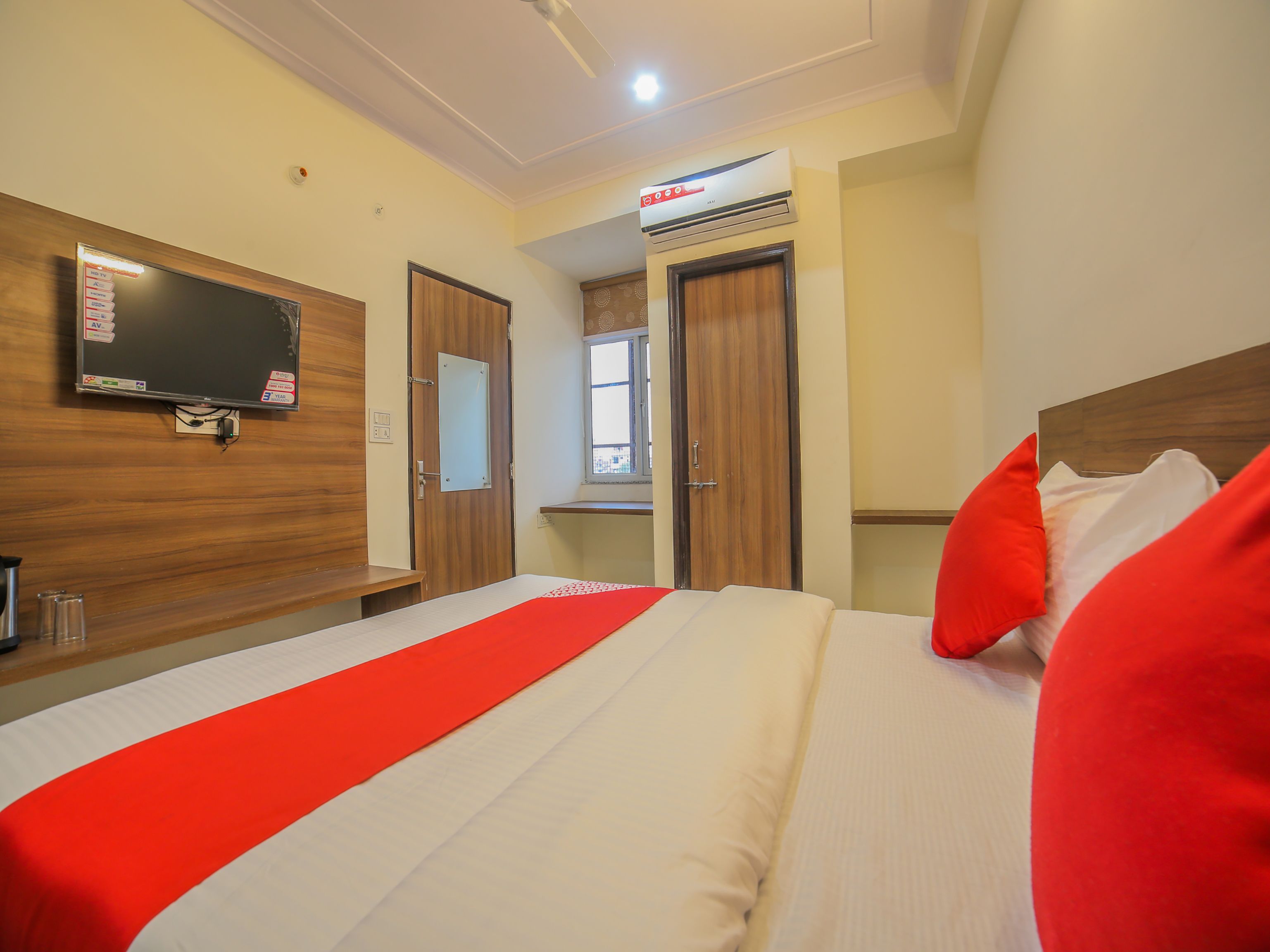 A hotel room with a bed, a TV mounted on the wall, a window, and an air conditioning unit.