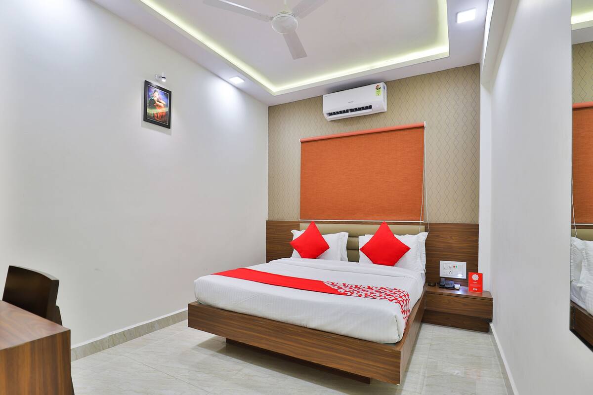 A modern hotel room with a wooden bed, red pillows, a ceiling fan, an air conditioning unit, and a small bedside table with a phone and a red sign.