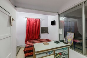 A small, sparsely furnished room with a bed, a table with books and a framed picture, a window with a red curtain, and a wall-mounted TV.