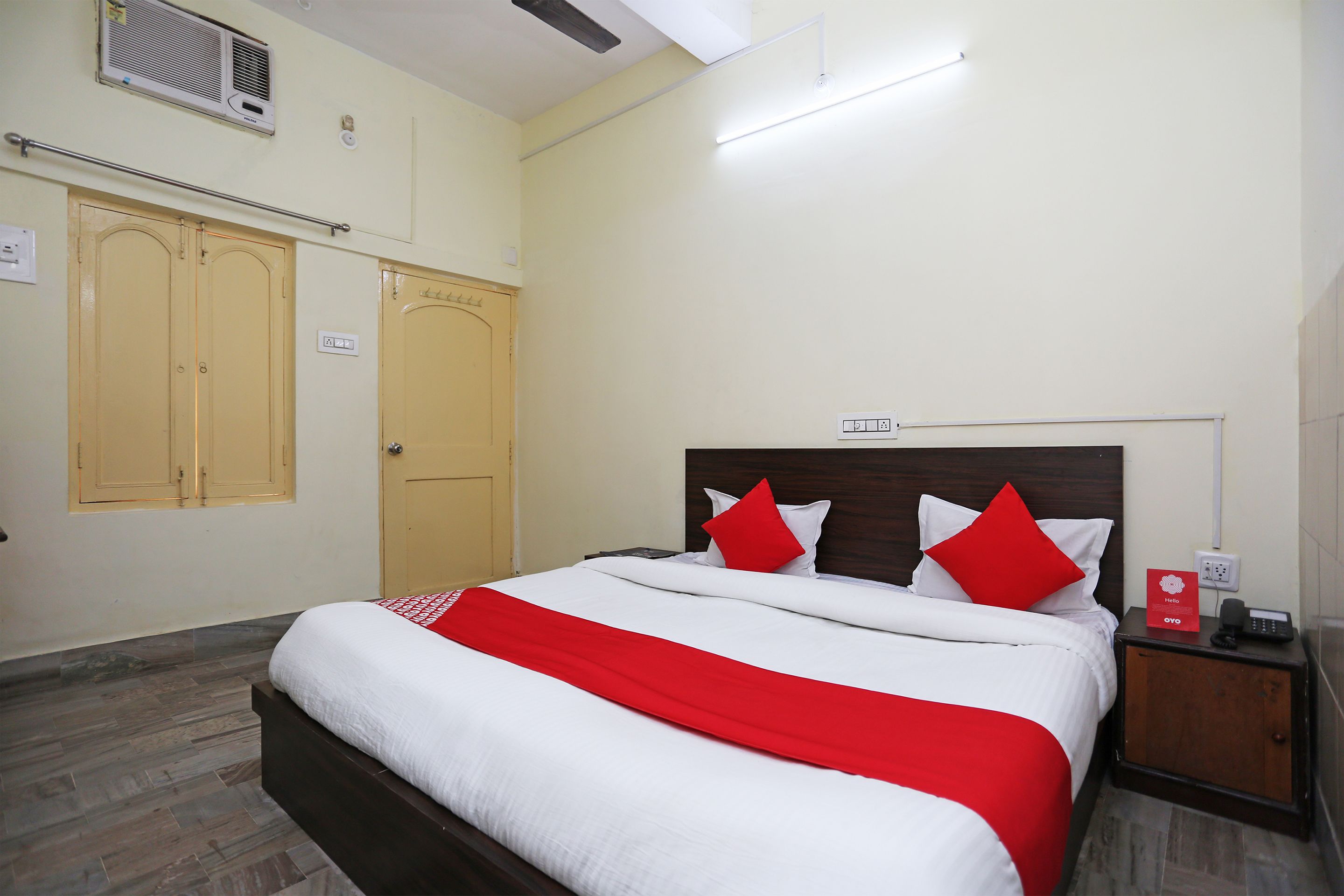 Habitación de hotel con cama, mesita de noche, cabecera de madera, colcha roja y blanca, y armario de madera.