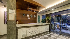 A hotel reception area with a marble counter, a clock displaying four time zones, and a sign stating no foreign currency exchange. - Capital O 28618 New Haven Hotel (New Delhi)