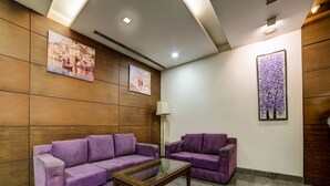 A room with two purple sofas, a glass coffee table, wooden panel walls, and artwork. - Capital O 28618 New Haven Hotel (New Delhi)