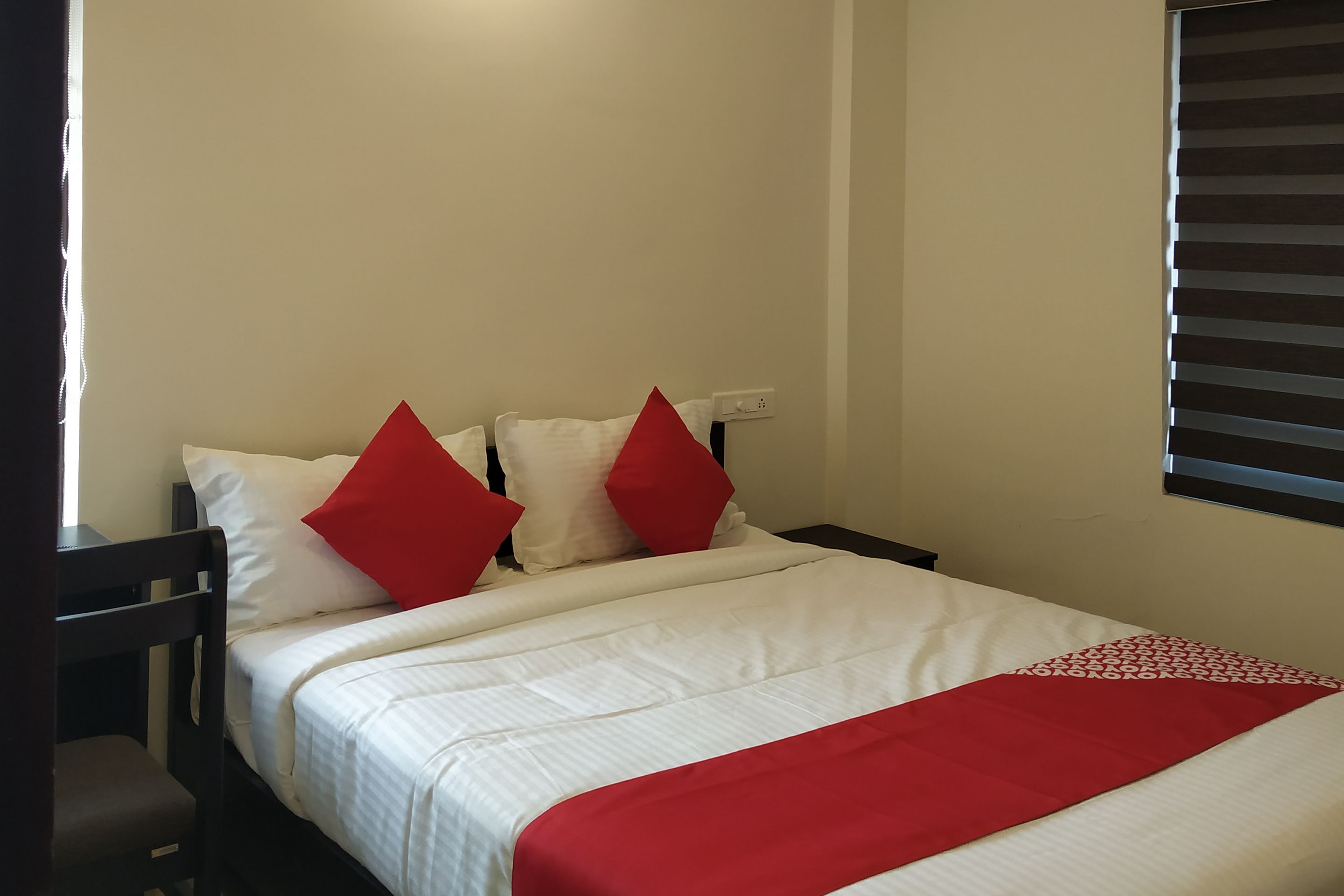 Una cama bien hecha con ropa de cama blanca y roja, una mesita de noche de madera y una ventana con persianas.