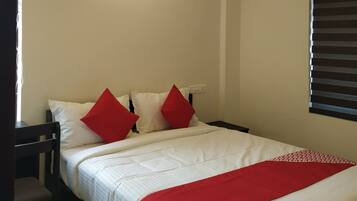 Una cama bien hecha con ropa de cama blanca y roja, una mesita de noche de madera y una ventana con persianas.