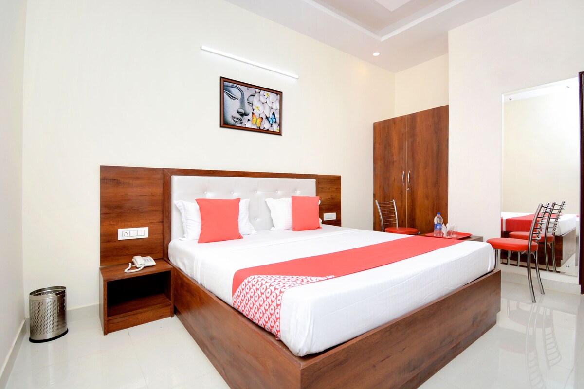 A modern hotel room with a large bed, wooden headboard, and a red and white bedspread. There is a small bedside table with a phone and a trash can. A painting hangs on the wall above the bed.