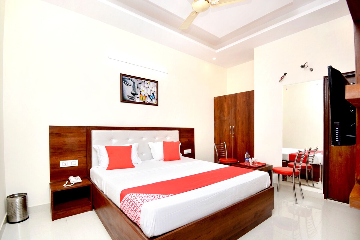A hotel room with a large bed, a wooden headboard, a red and white bedspread, a small bedside table with a phone, a trash can, and a mirror with a red chair.