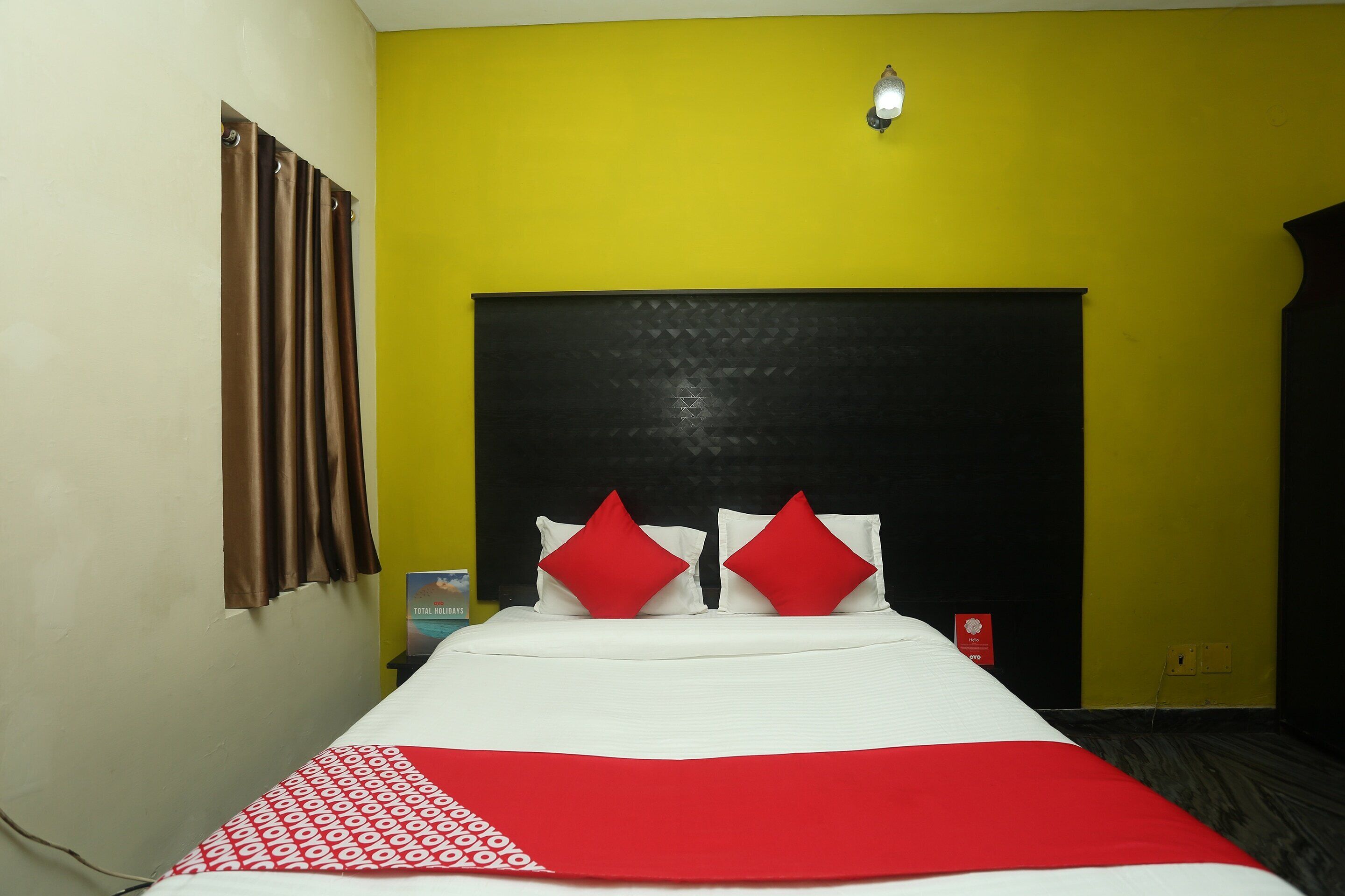 A hotel room with a bed, a headboard, a red and white bedspread, and a yellow wall.