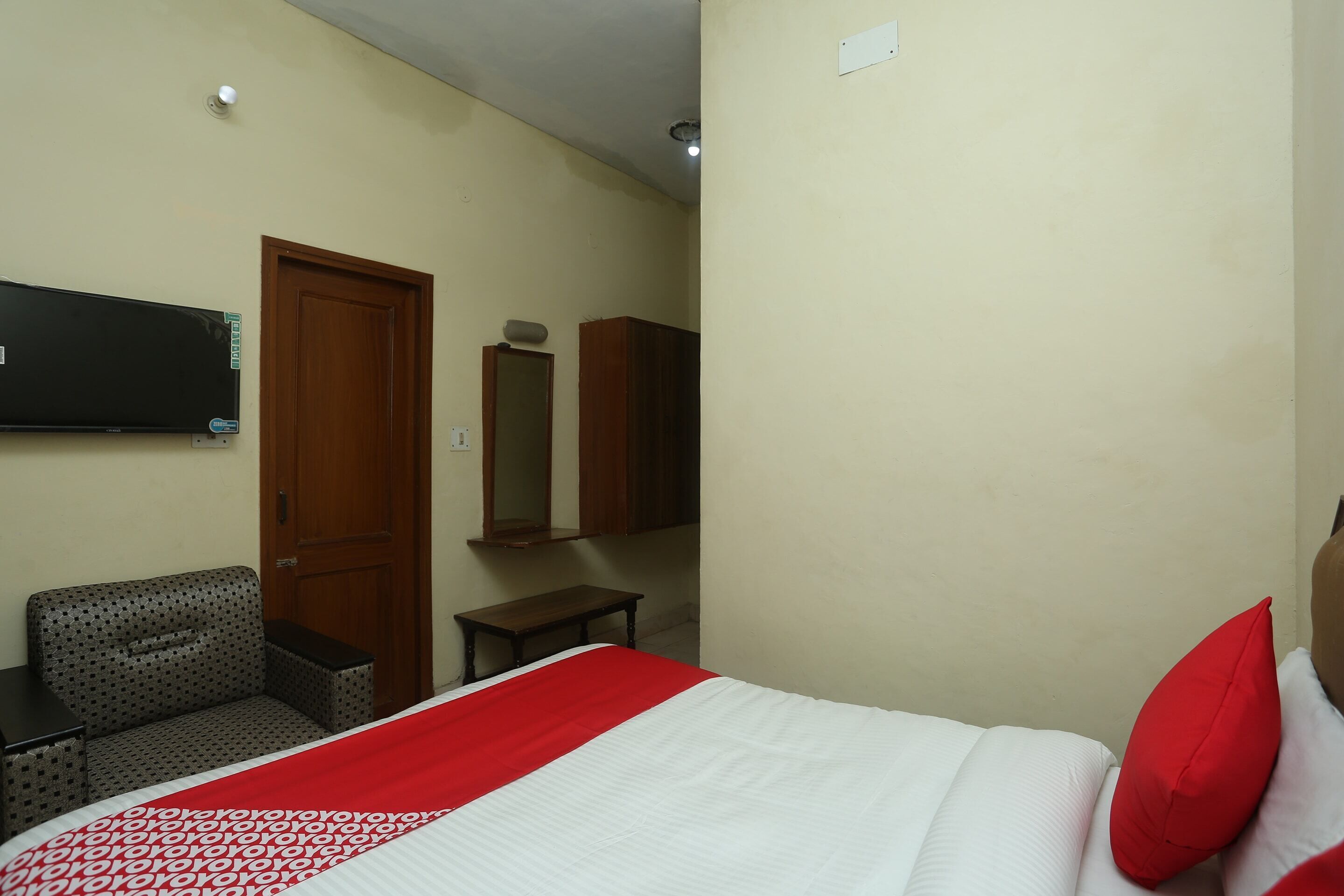 A hotel room with a bed, a TV mounted on the wall, a wooden door, a small table, and a bench.