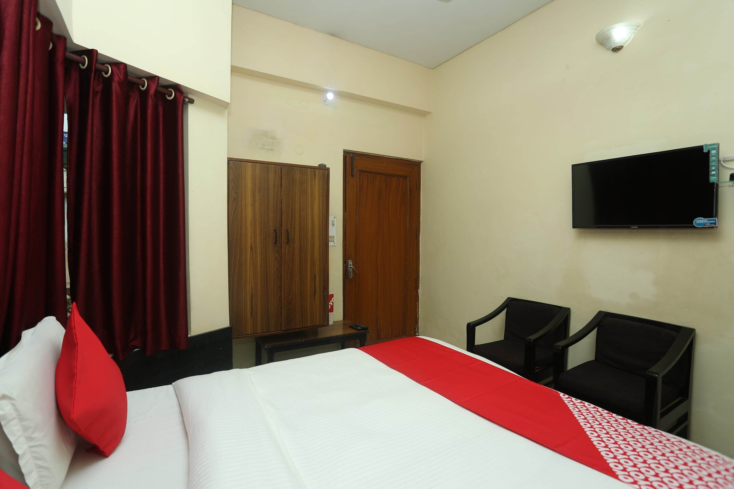 A hotel room with a bed, red curtains, a TV mounted on the wall, and two chairs.