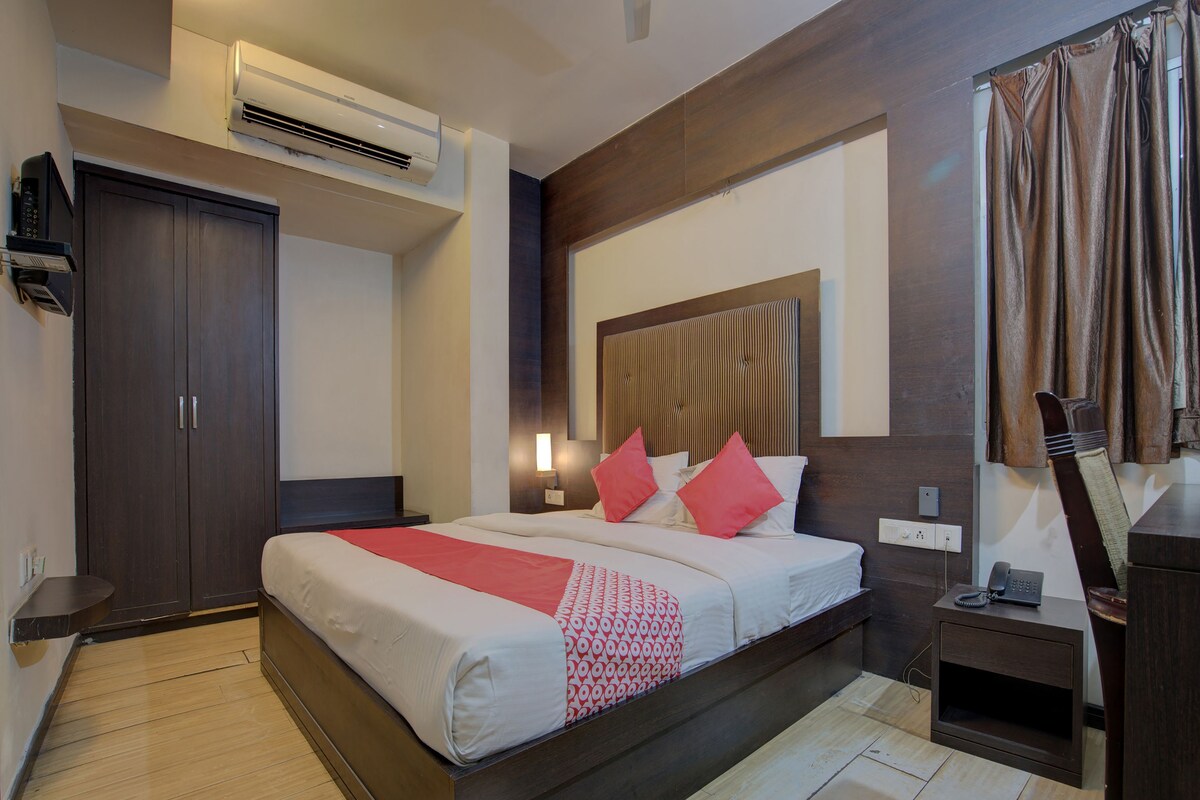 A hotel room with a large bed, a wardrobe, an air conditioning unit, a bedside table with a phone, and a small desk with a lamp.