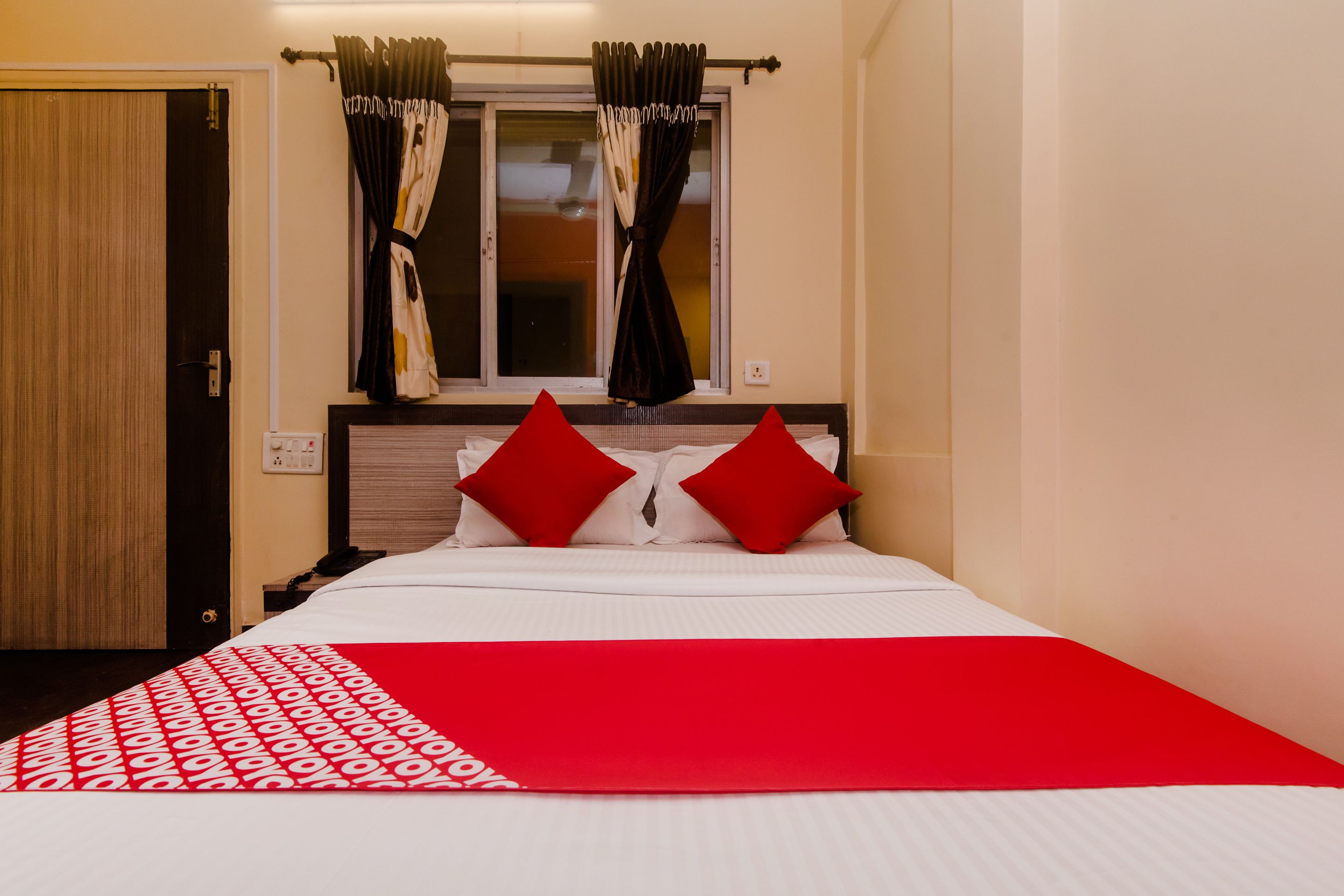 Habitación de hotel con cama, almohadones rojos, colcha con estampado rojo y blanco, y cortinas en una ventana.