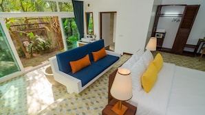 A bedroom with a bed, a blue sofa with orange cushions, a bedside table with a lamp, and a view of a garden through a glass door. - Draco Residence (Siem Reap)