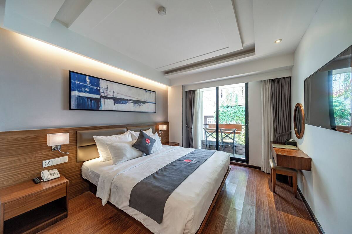 Ein Hotelzimmer mit einem großen Bett, Nachttischlampen, einem Telefon, einem Schreibtisch mit Stuhl und einem großen Fenster mit Vorhängen.