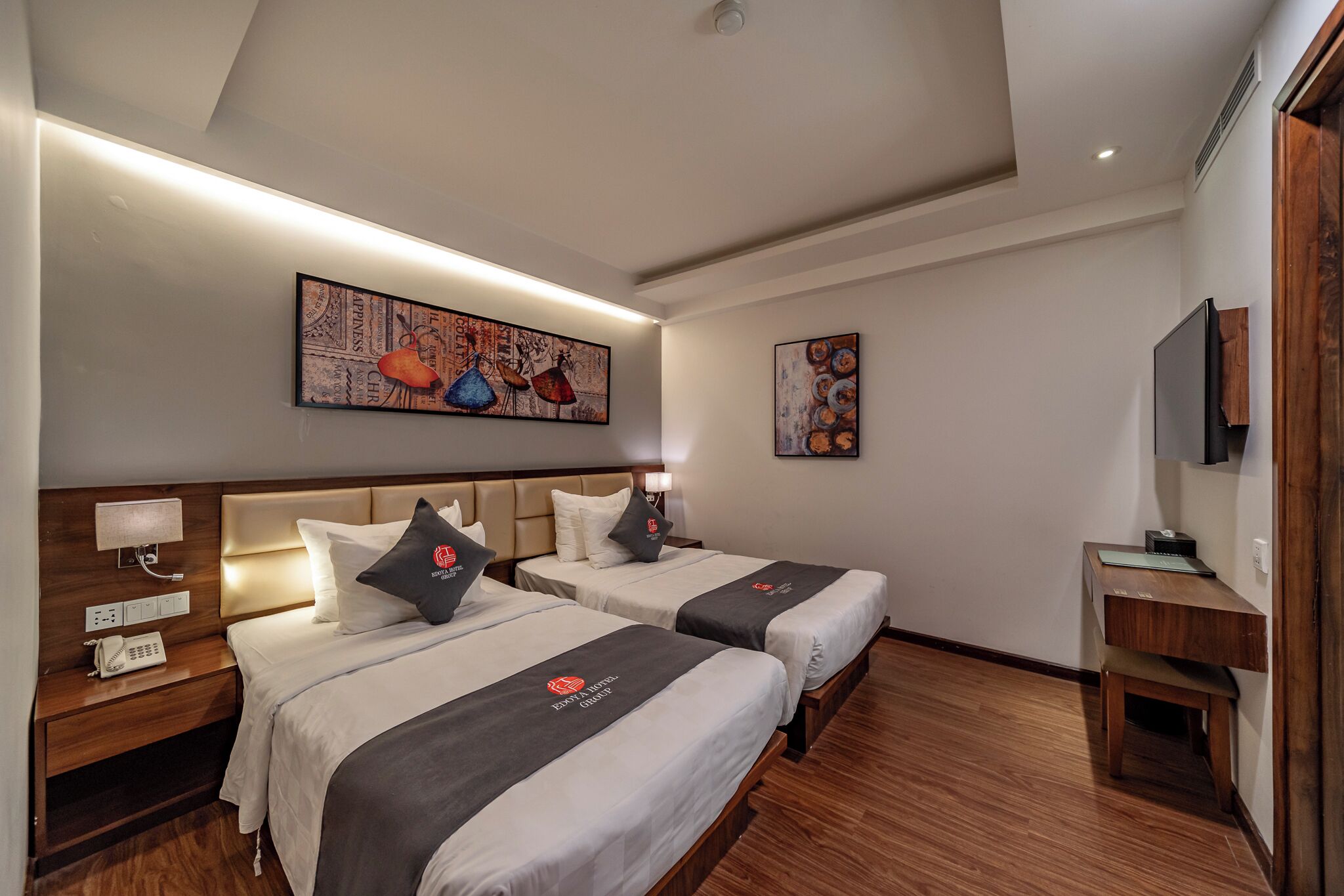 A hotel room with two beds, a wooden floor, a desk with a chair, a telephone, and artwork on the walls.