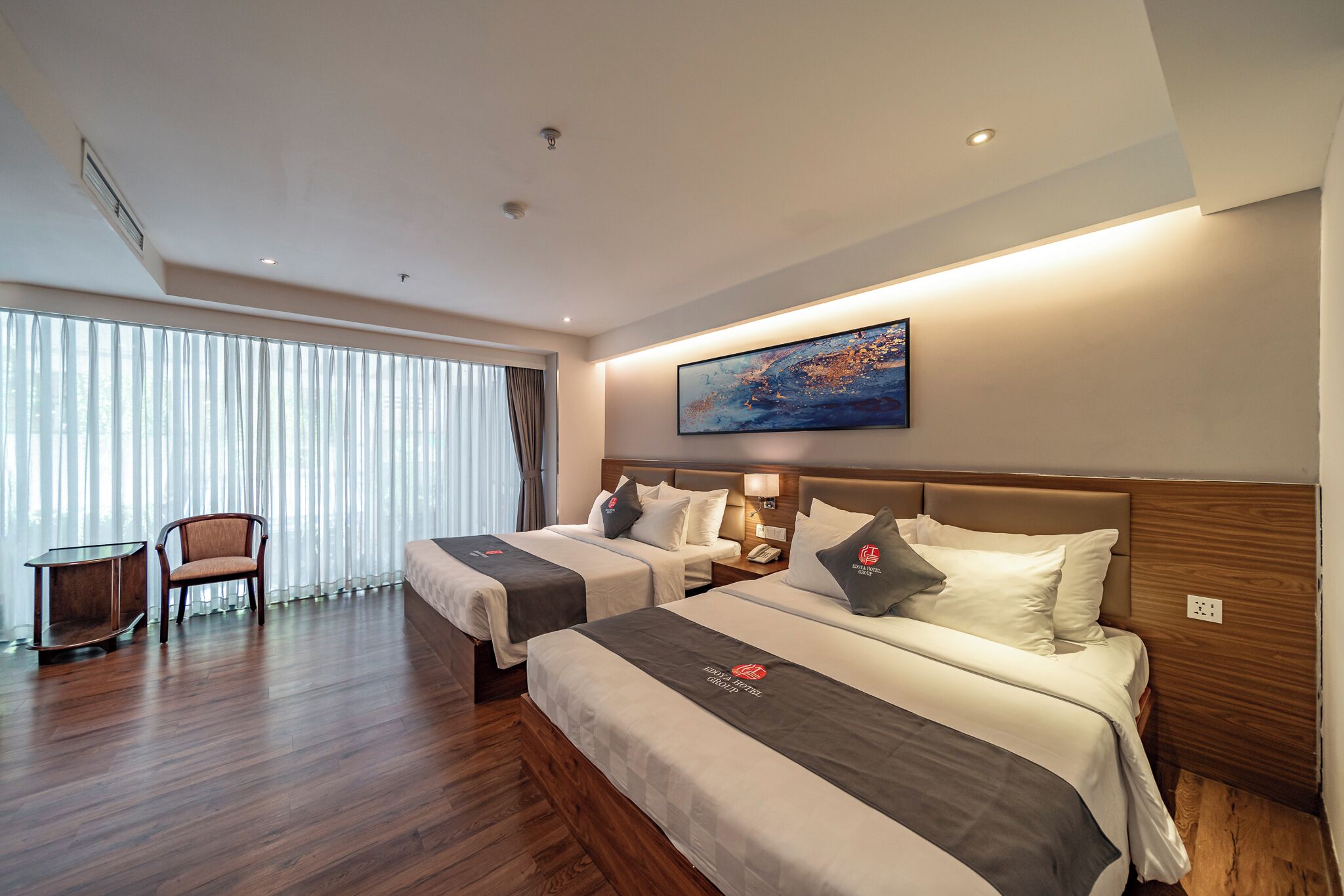 A hotel room with two beds, a small table, a chair, and a large window with curtains.