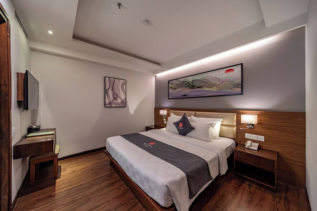 Ein modernes Hotelzimmer mit einem großen Bett, einem Holzkopfteil, Nachttischlampen, einem Schreibtisch mit Telefon und Kunstwerken an den Wänden.