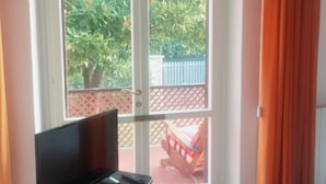 Une pièce dotée d’une porte-fenêtre coulissante donnant sur un balcon, d’un meuble de rangement noir pour équipements audiovisuels et d’un plancher carrelé.