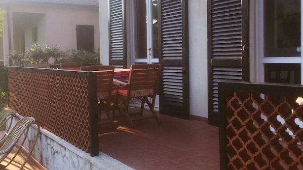 Une terrasse extérieure couverte, avec une table et des chaises, une clôture en bois et un bâtiment doté de grandes fenêtres et de volets.