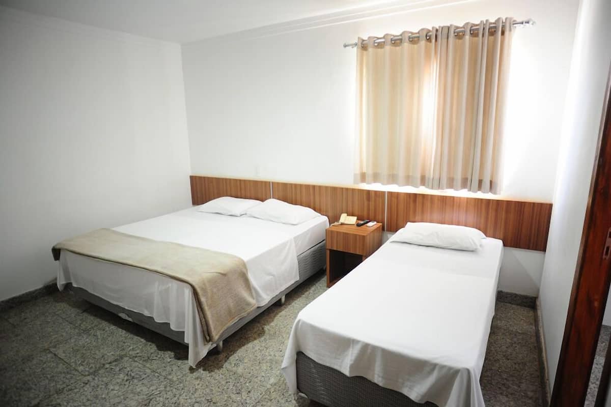 A hotel room with two single beds, a wooden headboard, a small bedside table with a lamp, and a window with curtains.