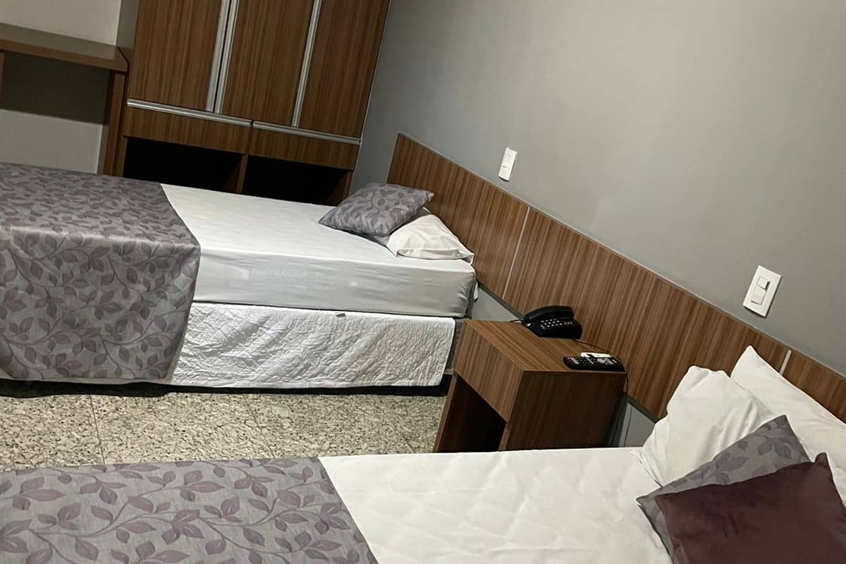 A hotel room with two beds, a wooden wardrobe, a bedside table with a phone, and a wall socket.