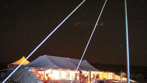 Un área con carpas, vista despejada del cielo nocturno, suelo arenoso y una silla de metal.