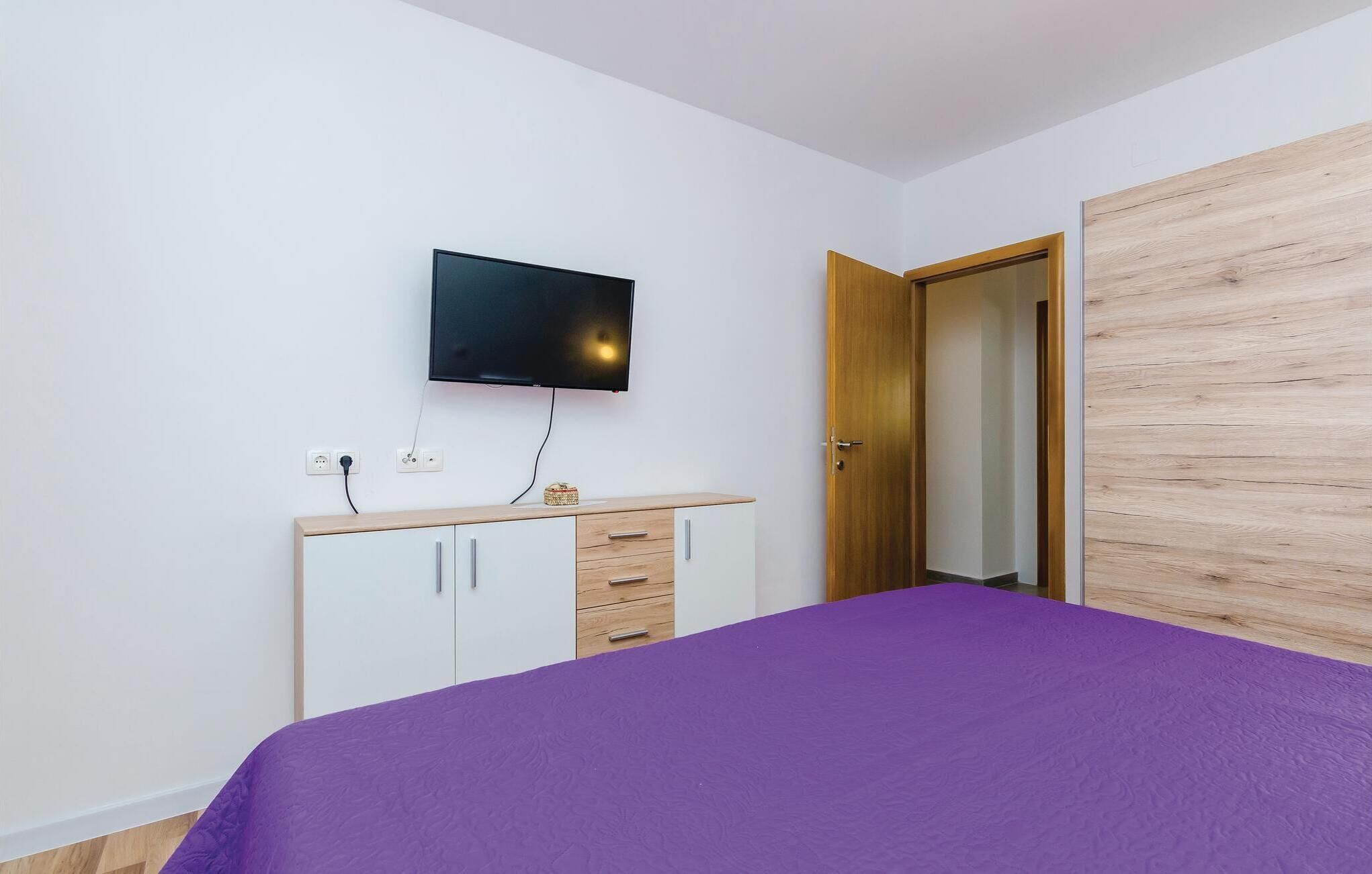 Une chambre à coucher comprenant un lit, un téléviseur fixé au mur, une armoire en bois et une porte donnant sur une autre pièce.