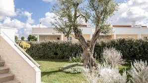 Un edificio residencial con una escalera que conduce a un balcón, rodeado de un jardín bien cuidado con arbustos variados y un árbol grande.
