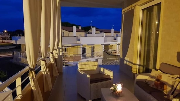 Un balcón con mobiliario de mimbre, una mesa con vela y vistas a edificios al fondo.