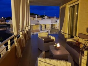 Un balcón con mobiliario de mimbre, una mesa con vela y vistas a edificios al fondo.