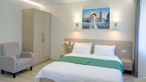A hotel room with a bed, a chair, a wardrobe, and a painting on the wall.