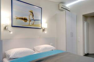 Una habitación de hotel con cama, paredes blancas, un cuadro enmarcado y un equipo de aire acondicionado.