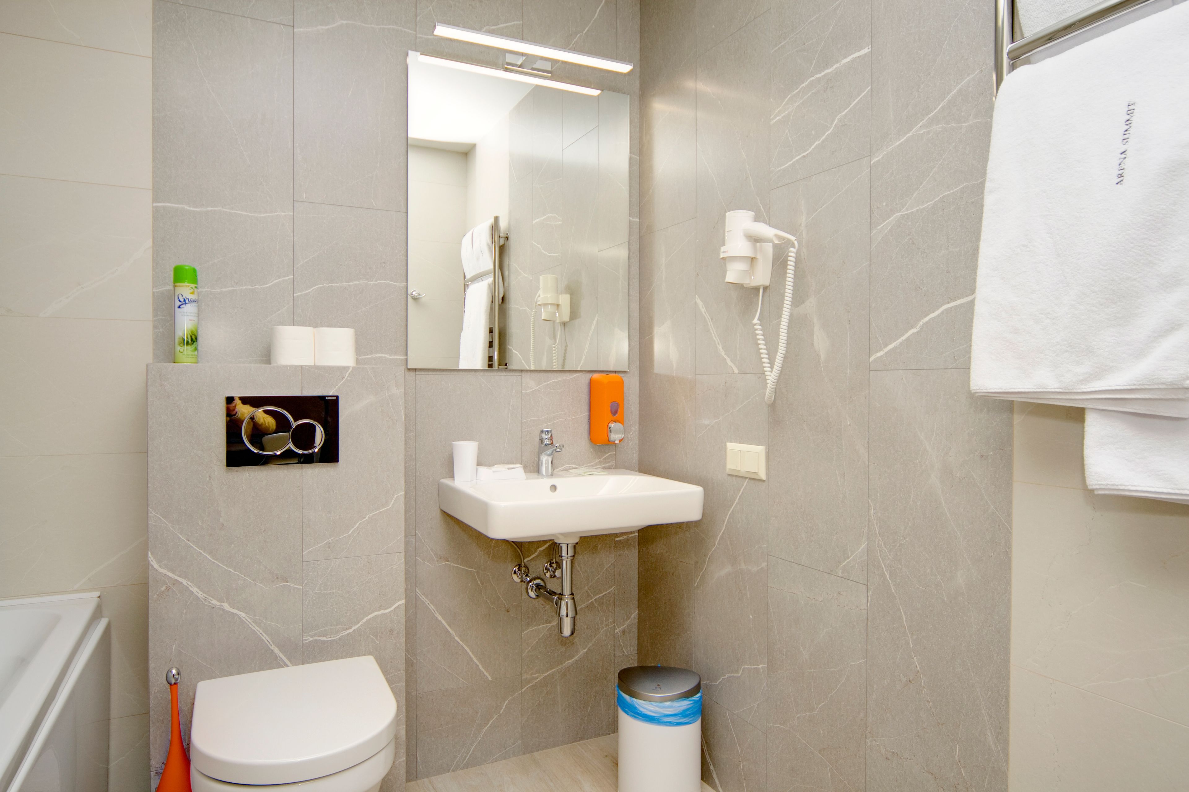 A bathroom with a toilet, sink, mirror, hairdryer, and a trash bin.