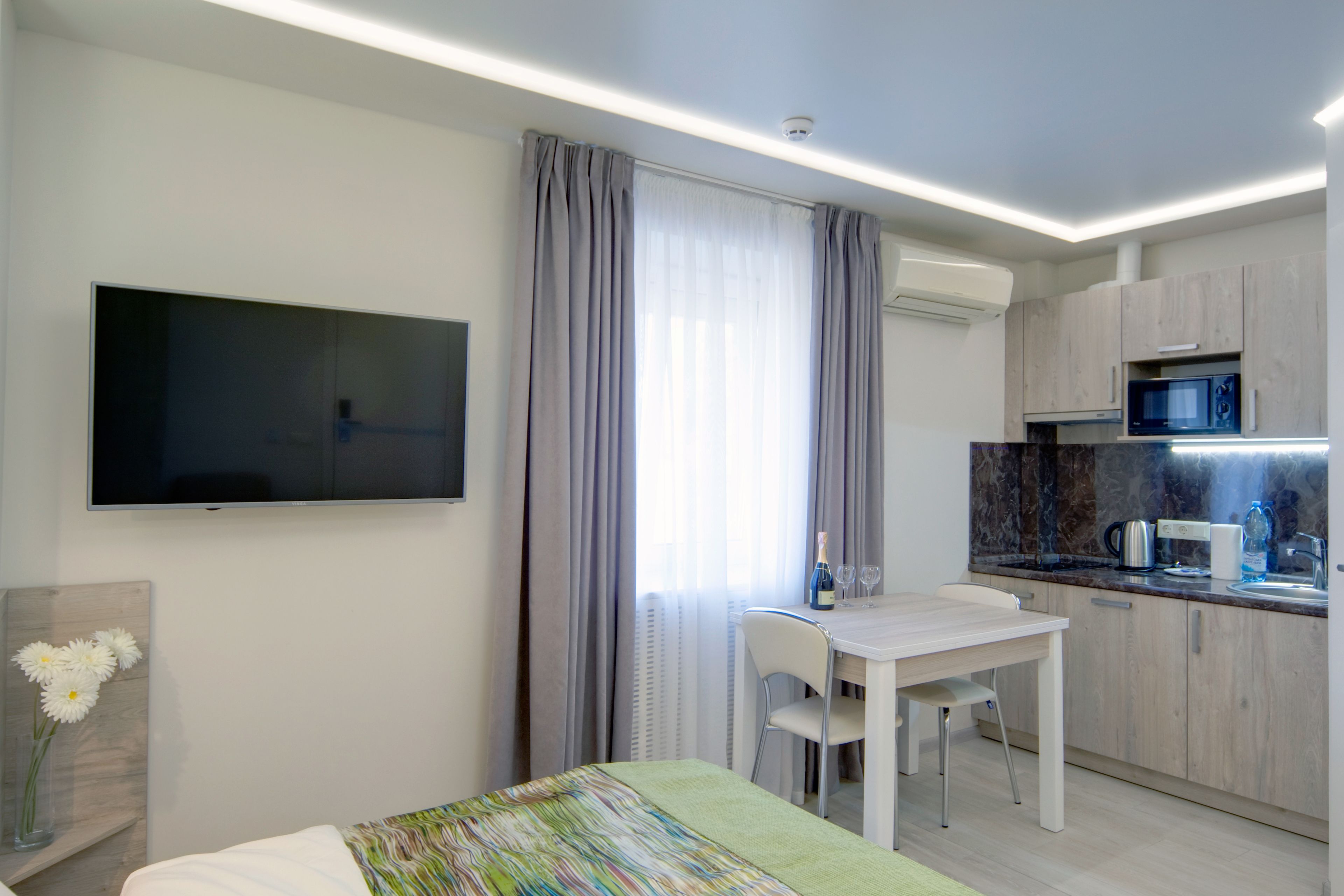 A modern hotel room with a bed, a TV, a dining table with chairs, a kitchen area with a microwave, and a large window with curtains.
