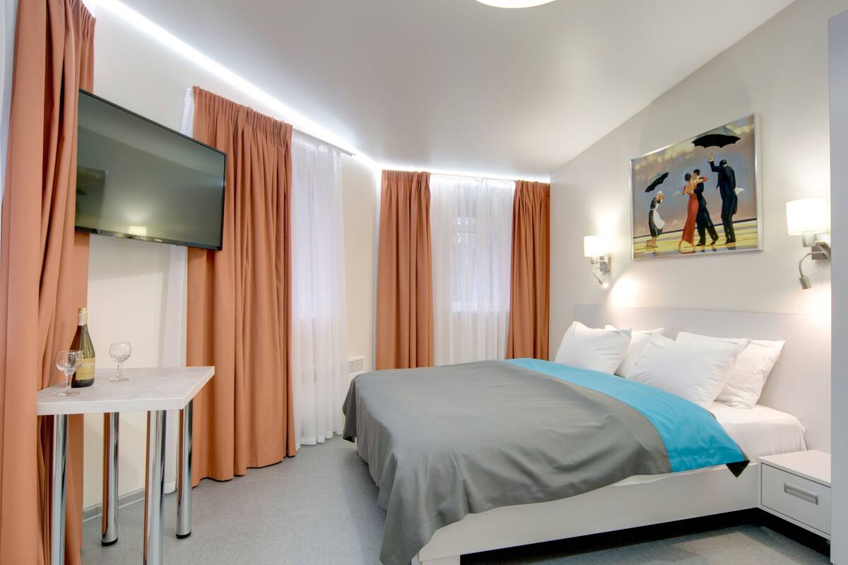 Ein Hotelzimmer mit Bett, einem an der Wand befestigten Fernseher, einem Tisch mit Flasche und Gläsern sowie einem Gemälde von einem Tanzpaar.