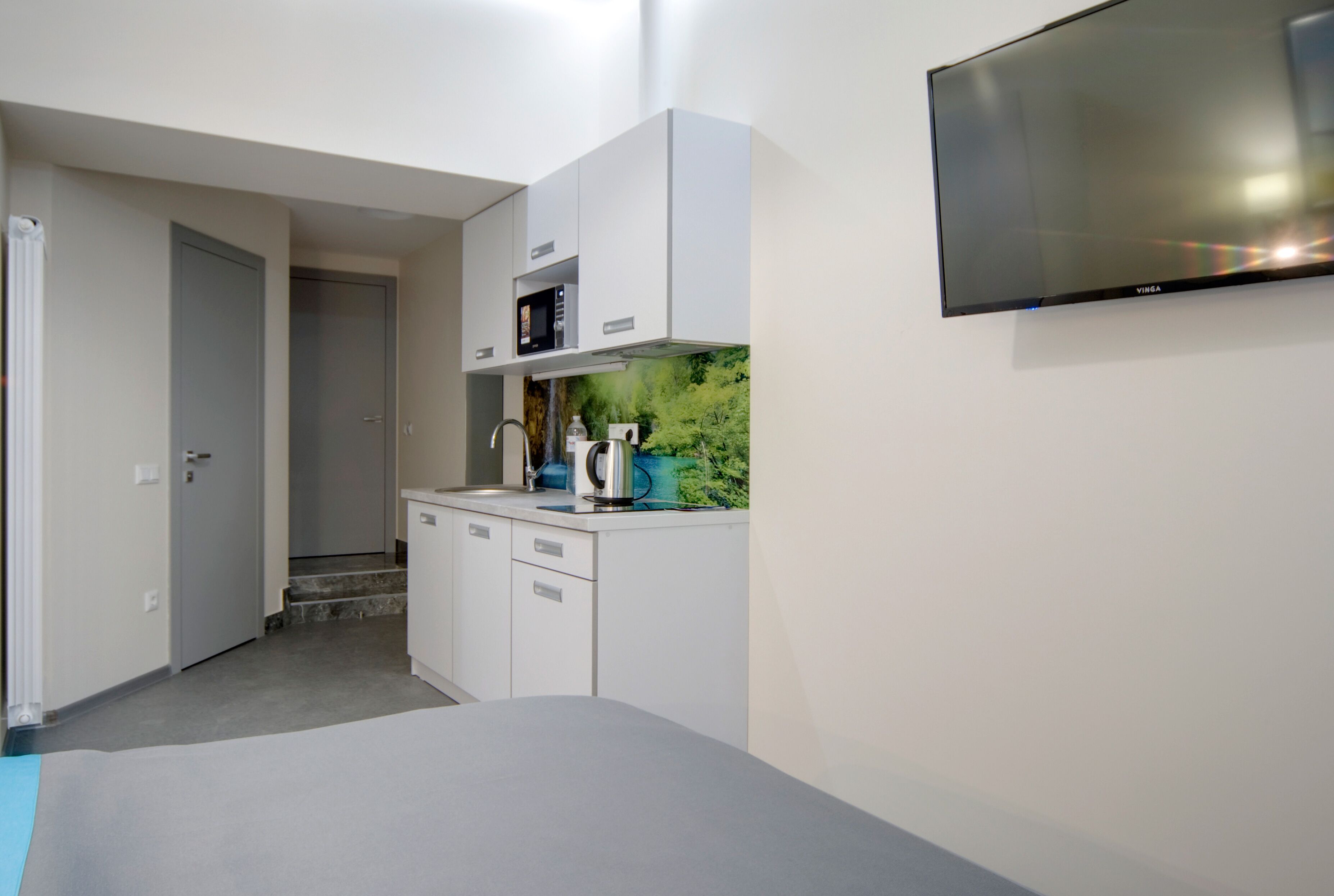 A compact room with a kitchenette, a bed, and a TV mounted on the wall.