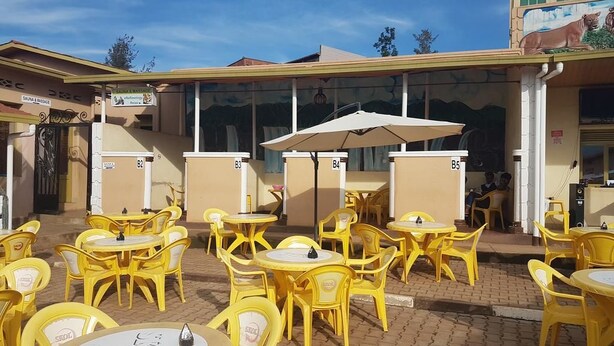 An outdoor seating area with yellow chairs and tables, a covered section with umbrellas, and a building with a mural. - Peace & Grace Motel (Kigali)