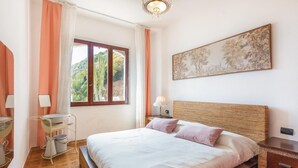 A bedroom with a bed, a bedside table, a lamp, a painting on the wall, and a window with curtains. - Stunning apartment in Porto Palma (Porto Palma)