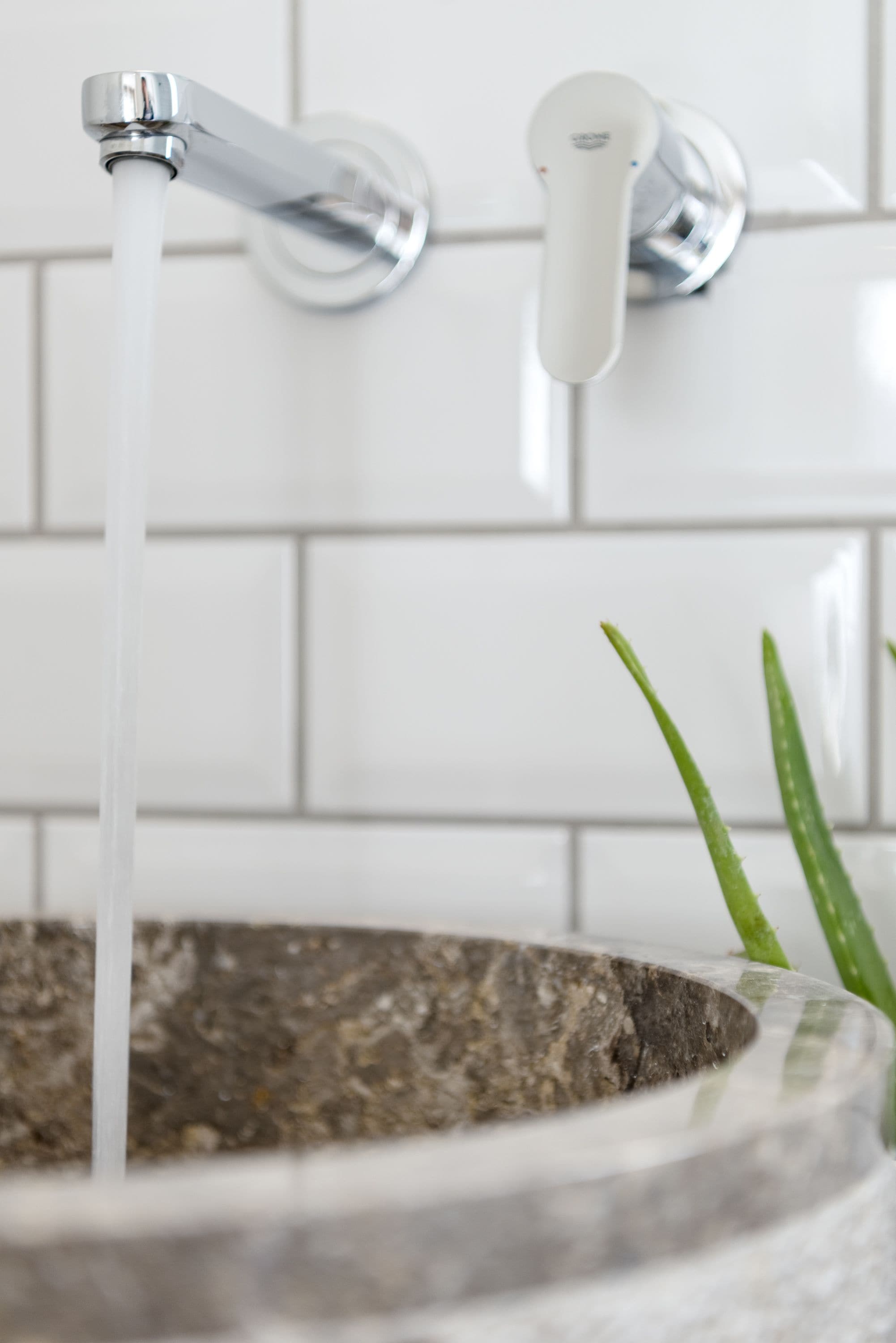 A bathroom with a white tiled wall, a chrome faucet, and a stone basin with a plant.