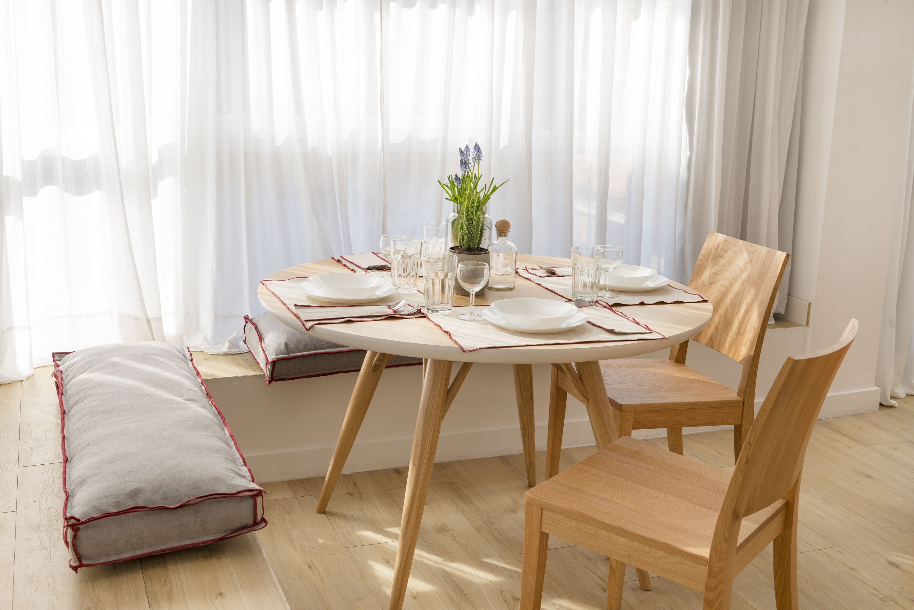 Un coin repas avec une table ronde dressée pour quatre personnes, des chaises en bois, un banc et une fenêtre ornée de rideaux légers.