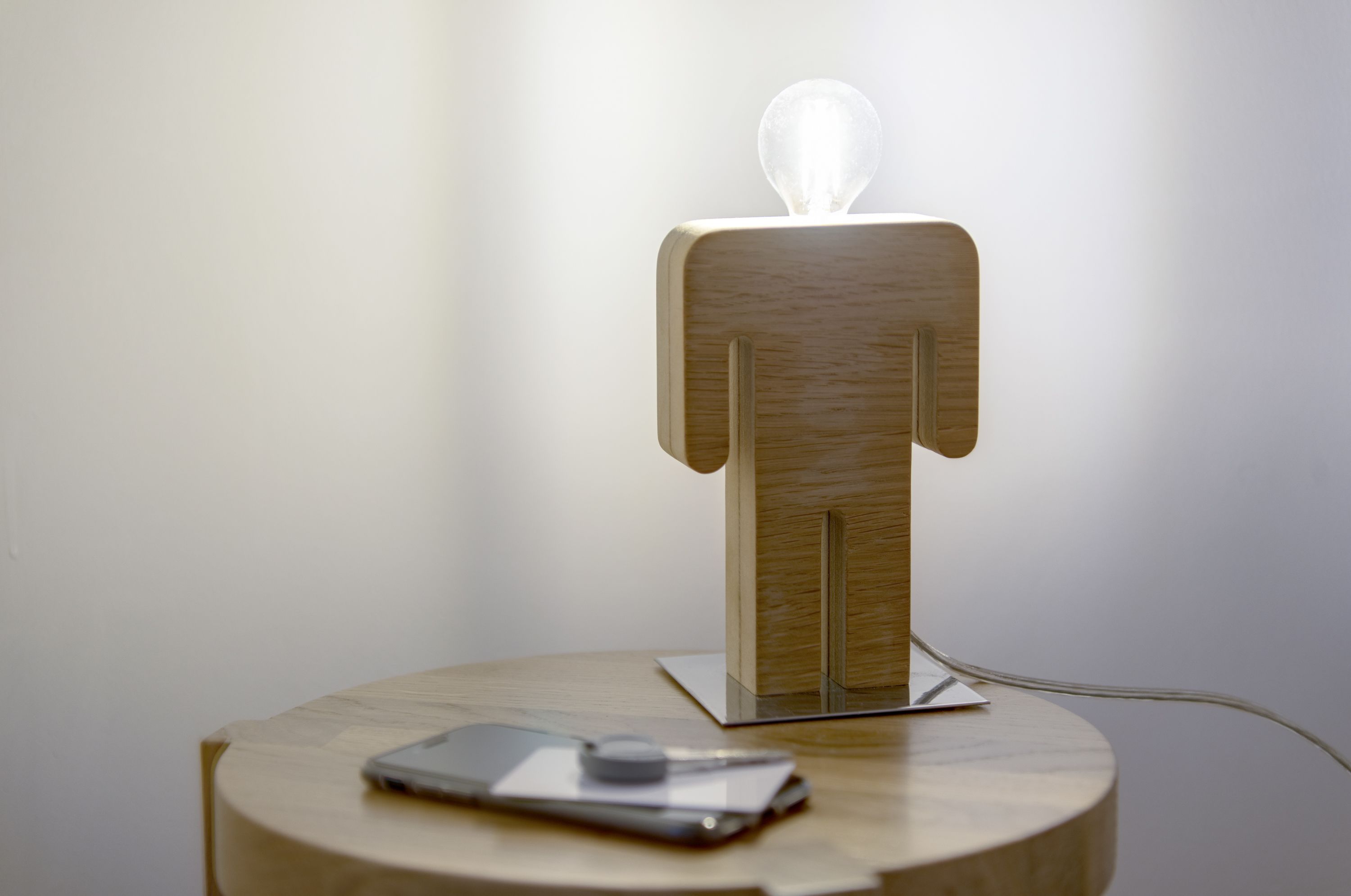 Una figura de madera con una bombilla en la cabeza, sobre una mesa redonda con un teléfono inteligente y un objeto pequeño.
