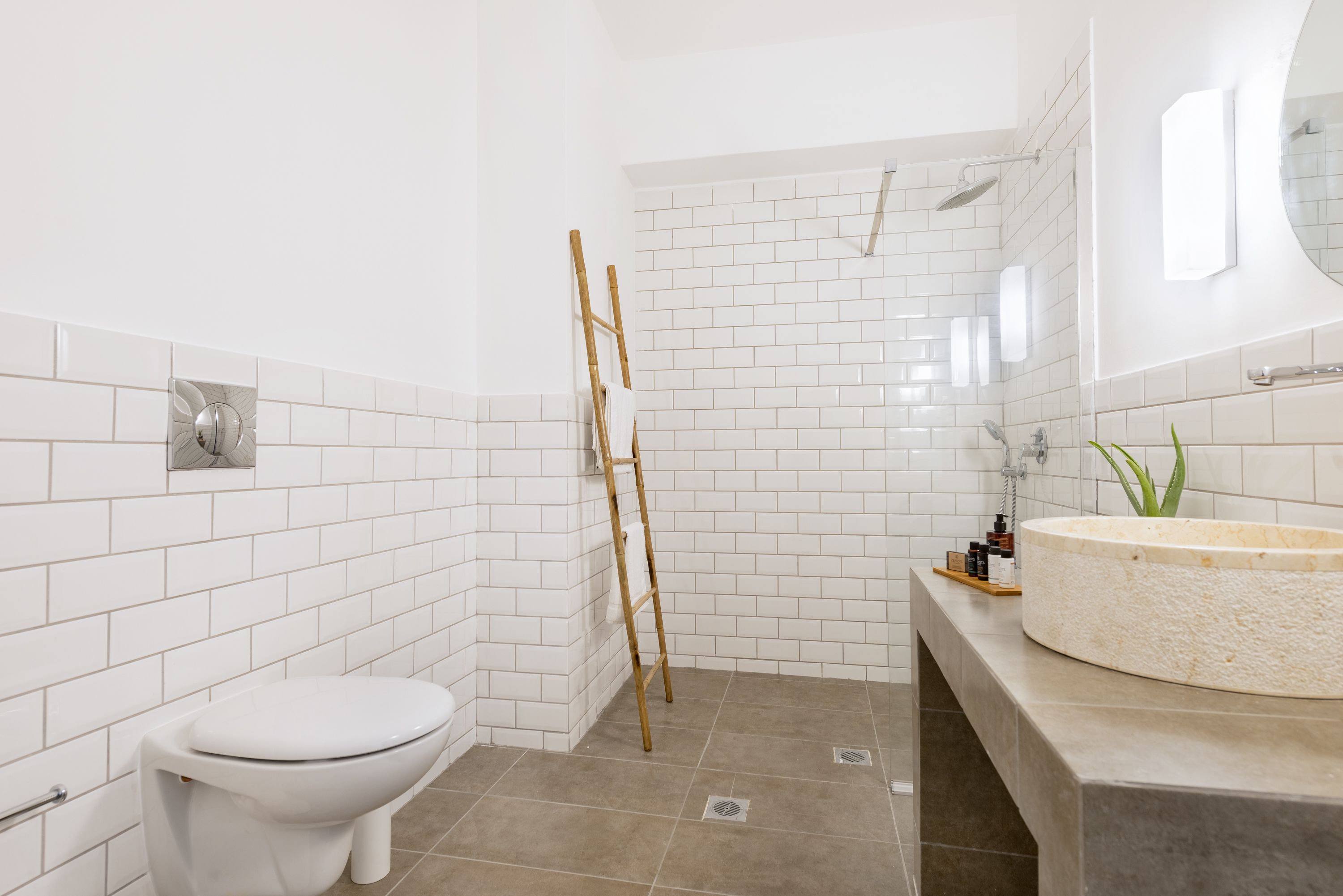 Ein Badezimmer mit weißen Fliesen, einer Toilette, einer Duschkabine und einem Waschtisch mit rundem Becken.