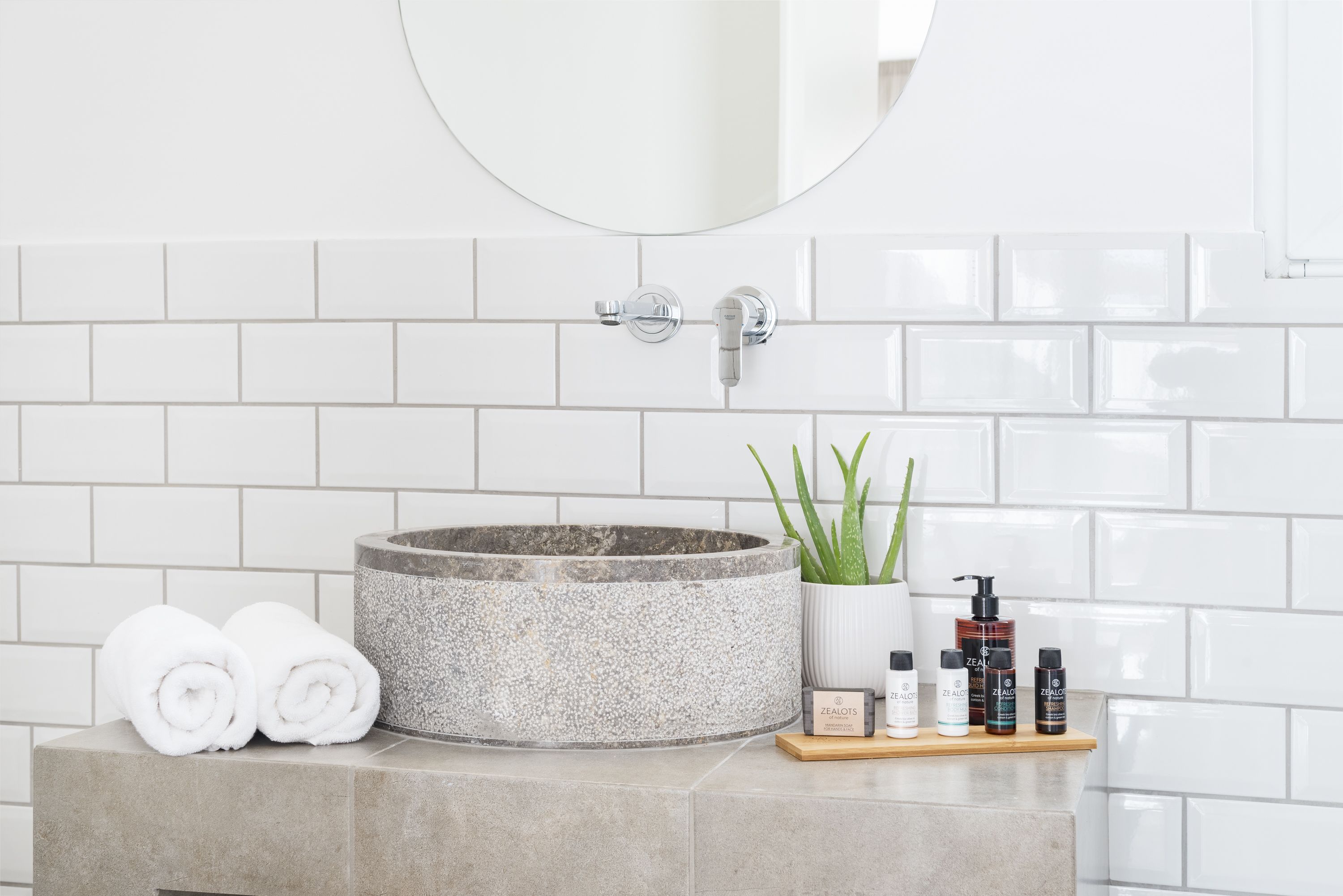 A bathroom with a round stone sink, white tiles, a mirror, a potted aloe vera plant, and neatly folded towels.