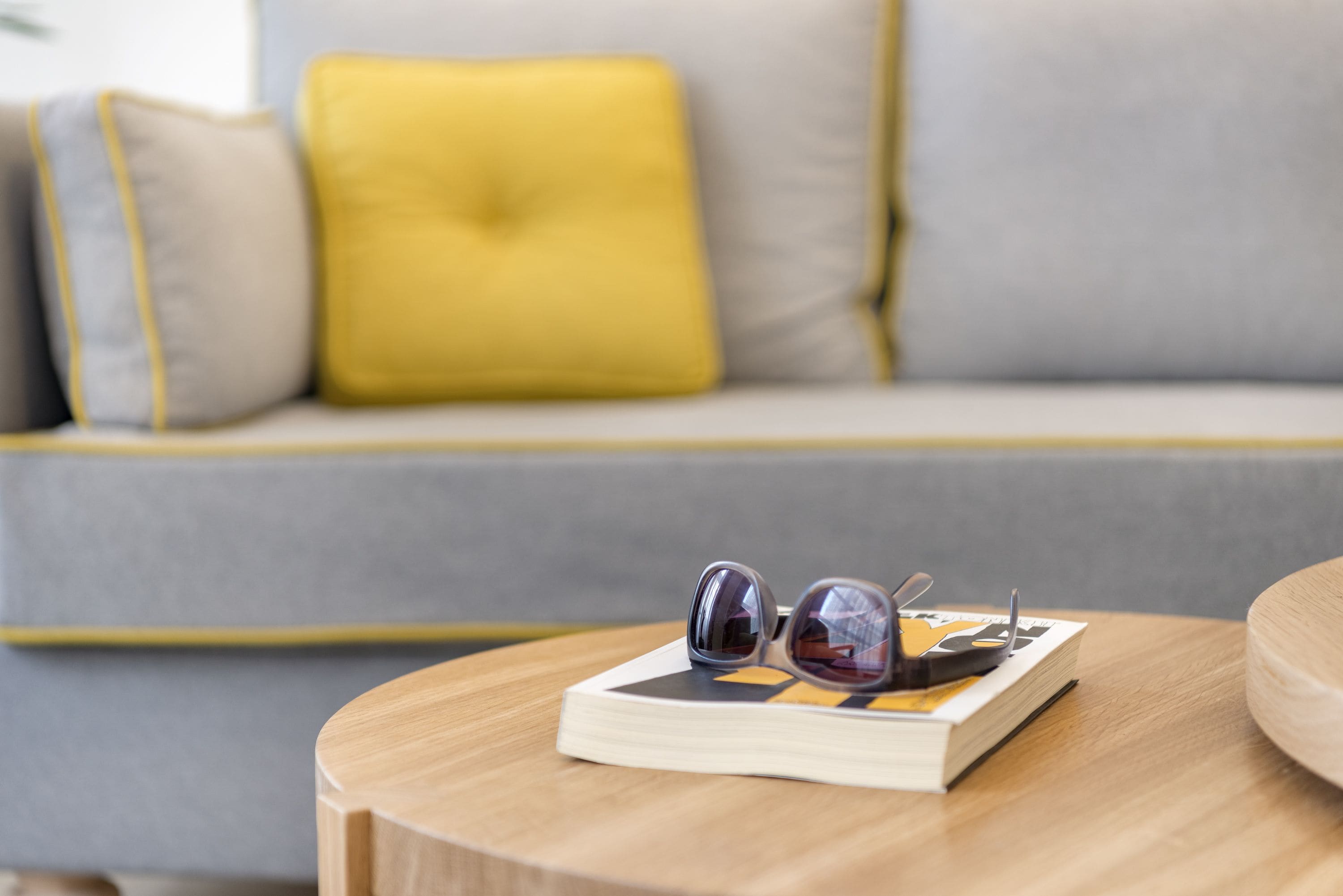 Une paire de lunettes de soleil posée sur un livre, lui-même posé sur une table en bois. On aperçoit un canapé et des coussins à l’arrière-plan.